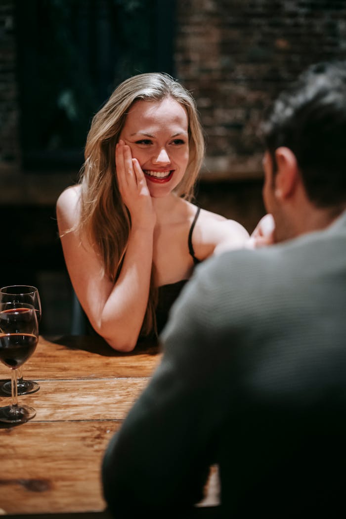 Young happy couple in elegant wear sitting at table in restaurant and enjoying date while with red wine in glasses and kissing hand gently